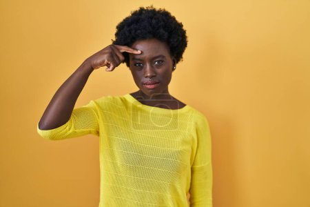 Foto de Mujer joven africana de pie sobre el estudio amarillo señalando infeliz a la espinilla en la frente, fea infección de la cabeza negra. acné y problemas de piel - Imagen libre de derechos