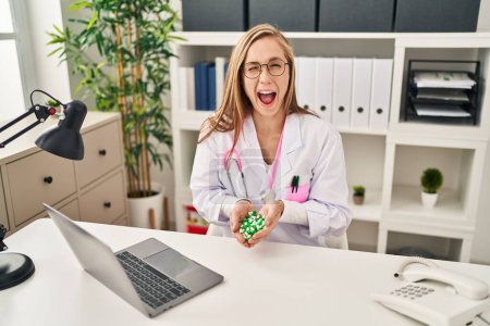 Téléchargez les photos : Jeune femme blonde médecin tenant des pilules clin d'oeil en regardant la caméra avec une expression sexy, gai et heureux visage. - en image libre de droit