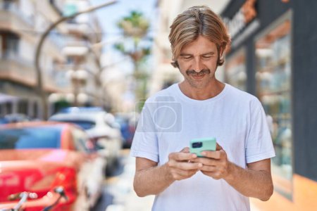 Téléchargez les photos : Jeune homme blond souriant confiant en utilisant un smartphone dans la rue - en image libre de droit