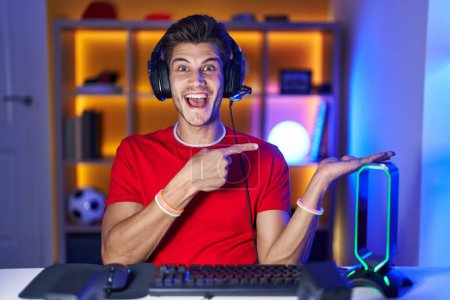 Photo for Young hispanic man playing video games amazed and smiling to the camera while presenting with hand and pointing with finger. - Royalty Free Image