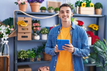 Téléchargez les photos : Jeune homme hispanique fleuriste souriant confiant en utilisant le pavé tactile au magasin de fleuriste - en image libre de droit