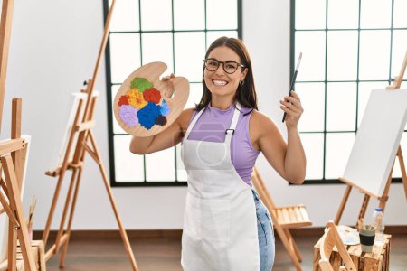 Foto de Joven hermosa artista hispana sonriendo confiada sosteniendo pincel y paleta en el estudio de arte - Imagen libre de derechos