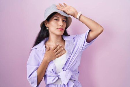 Téléchargez les photos : Jeune femme hispanique debout sur fond rose portant un chapeau touchant le front pour la maladie et la fièvre, la grippe et le rhume, le virus malade - en image libre de droit