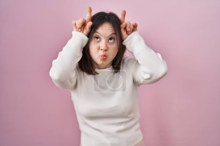 Téléchargez les photos : Femme avec le syndrome du duvet debout sur fond rose faisant un geste drôle avec le doigt sur la tête comme des cornes de taureau - en image libre de droit
