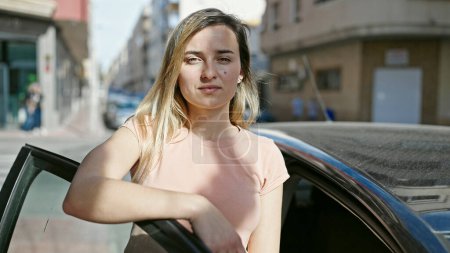 Photo for Young blonde woman leaning on car door with relaxed expression at street - Royalty Free Image