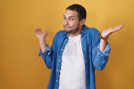 Photo for Hispanic man standing over yellow background clueless and confused expression with arms and hands raised. doubt concept. - Royalty Free Image