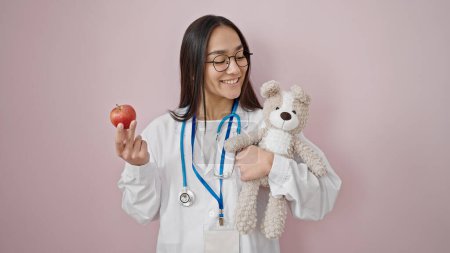 Photo for Young beautiful hispanic woman doctor smiling confident holding teddy bear and apple over isolated pink background - Royalty Free Image