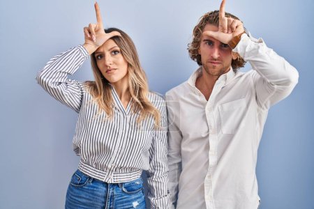 Photo for Young couple standing over blue background making fun of people with fingers on forehead doing loser gesture mocking and insulting. - Royalty Free Image
