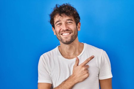 Téléchargez les photos : Jeune homme hispanique debout sur fond bleu joyeux avec un sourire sur le visage pointant avec la main et le doigt vers le côté avec une expression heureuse et naturelle - en image libre de droit