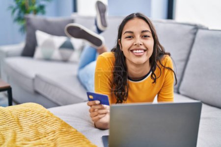 Téléchargez les photos : Jeune femme afro-américaine utilisant un ordinateur portable et une carte de crédit couchée sur le canapé à la maison - en image libre de droit