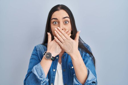 Photo for Hispanic woman standing over blue background shocked covering mouth with hands for mistake. secret concept. - Royalty Free Image