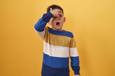 Foto de Pequeño niño hispano de pie sobre fondo amarillo asomándose en shock cubriendo la cara y los ojos con la mano, mirando a través de los dedos asustado - Imagen libre de derechos