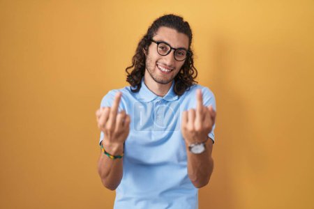 Foto de Young hispanic man standing over yellow background showing middle finger doing fuck you bad expression, provocation and rude attitude. screaming excited - Imagen libre de derechos