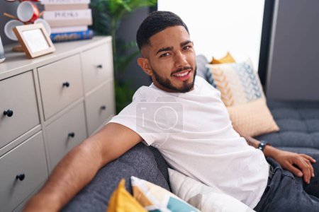 Téléchargez les photos : Jeune homme latino souriant confiant assis sur le canapé à la maison - en image libre de droit