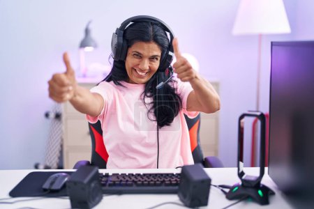 Téléchargez les photos : Femme hispanique mature jouant à des jeux vidéo à la maison approuvant faire un geste positif avec la main, pouces levés souriant et heureux pour le succès. geste gagnant. - en image libre de droit