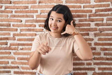 Foto de Mujer hispana joven de pie sobre la pared de ladrillos sonriendo haciendo hablar por teléfono gesto y señalando a usted. Llámame.. - Imagen libre de derechos
