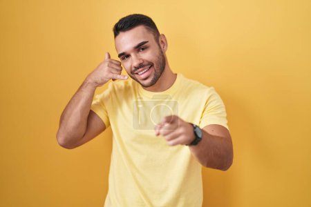 Téléchargez les photos : Jeune homme hispanique debout sur fond jaune souriant faisant parler au téléphone geste et vous pointant du doigt. Appelle-moi. - en image libre de droit