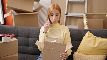Photo for Young blonde woman talking on smartphone unpacking cardboard box at new home - Royalty Free Image