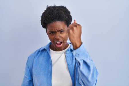 Foto de Mujer afroamericana de pie sobre fondo azul enojado y loco levantando el puño frustrado y furioso mientras gritaba con ira. rabia y concepto agresivo. - Imagen libre de derechos