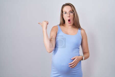 Photo for Young pregnant woman standing over white background surprised pointing with hand finger to the side, open mouth amazed expression. - Royalty Free Image