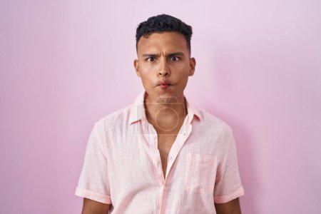 Foto de Young hispanic man standing over pink background making fish face with lips, crazy and comical gesture. funny expression. - Imagen libre de derechos
