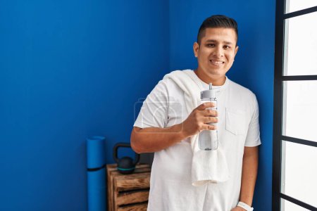 Téléchargez les photos : Jeune homme latino souriant confiant tenant bouteille d'eau au centre sportif - en image libre de droit