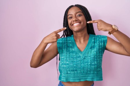 Téléchargez les photos : Jeune afro-américain avec des tresses debout sur fond rose souriant gai montrant et pointant avec les doigts les dents et la bouche. concept de santé dentaire. - en image libre de droit
