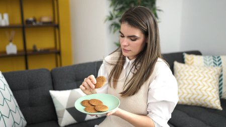 Foto de Joven mujer hispana hermosa comiendo galletas sentada en el sofá en casa - Imagen libre de derechos