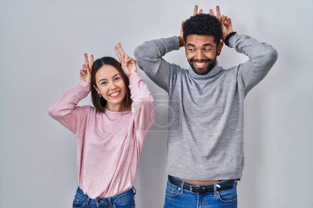 Foto de Joven pareja hispana de pie juntos posando divertida y loca con los dedos en la cabeza como orejas de conejo, sonriendo alegre - Imagen libre de derechos