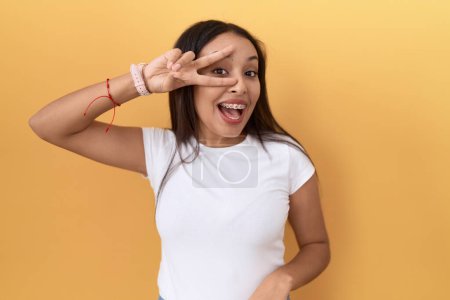 Téléchargez les photos : Jeune femme arabe portant un t-shirt blanc décontracté sur fond jaune faisant symbole de paix avec les doigts sur le visage, souriant joyeux montrant la victoire - en image libre de droit