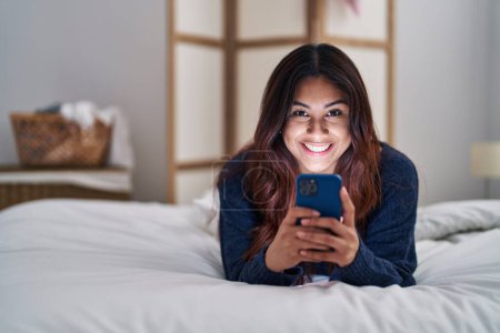 Téléchargez les photos : Young hispanic woman using smartphone lying on bed at bedroom - en image libre de droit