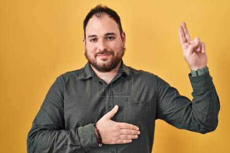 Foto de Hombre hispano de talla grande con barba de pie sobre fondo amarillo sonriendo jurando con la mano en el pecho y los dedos hacia arriba, haciendo un juramento de lealtad promesa - Imagen libre de derechos