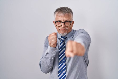 Foto de Hombre de negocios hispano con el pelo gris usando gafas golpeando puño para luchar, ataque agresivo y enojado, amenaza y violencia - Imagen libre de derechos