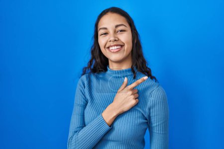 Photo for Young brazilian woman standing over blue isolated background cheerful with a smile on face pointing with hand and finger up to the side with happy and natural expression - Royalty Free Image