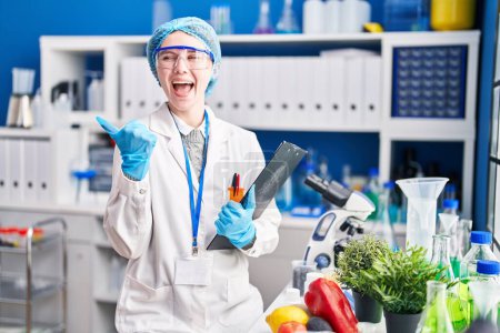 Foto de Hermosa mujer que trabaja en el laboratorio científico con comida apuntando el pulgar hacia el lado sonriendo feliz con la boca abierta - Imagen libre de derechos