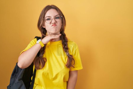 Téléchargez les photos : Jeune femme caucasienne portant un sac à dos étudiant sur fond jaune tranchant la gorge avec la main comme couteau, menace d'agression avec une violence furieuse - en image libre de droit