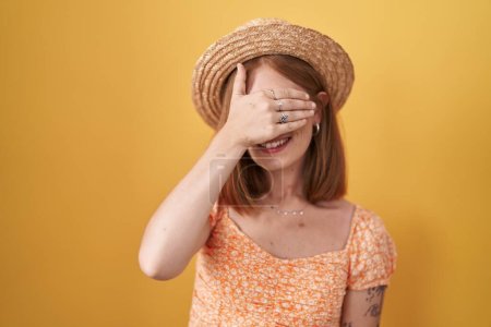 Photo for Young redhead woman standing over yellow background wearing summer hat smiling and laughing with hand on face covering eyes for surprise. blind concept. - Royalty Free Image