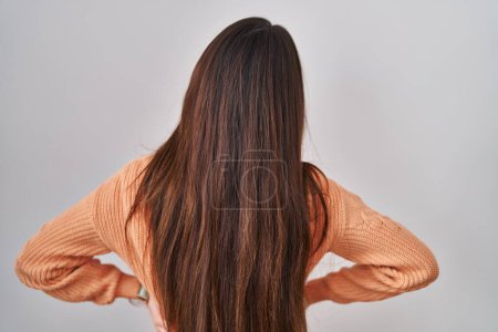 Téléchargez les photos : Jeune femme brune debout sur fond blanc regardant vers l'arrière avec les bras sur le corps - en image libre de droit