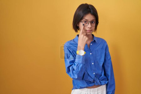 Téléchargez les photos : Jeune fille debout sur fond jaune pointant vers l'oeil vous regardant geste, expression suspecte - en image libre de droit