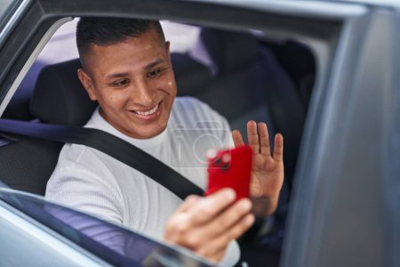 Foto de Joven hispano haciendo videollamada con smartphone en el coche luciendo positivo y feliz de pie y sonriendo con una sonrisa confiada mostrando los dientes - Imagen libre de derechos
