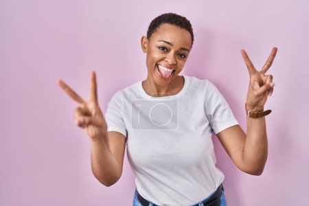 Téléchargez les photos : Belle femme afro-américaine debout sur fond rose souriant avec la langue montrant les doigts des deux mains faisant signe de victoire. numéro deux. - en image libre de droit