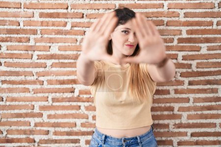 Photo for Young brunette woman standing over bricks wall doing frame using hands palms and fingers, camera perspective - Royalty Free Image
