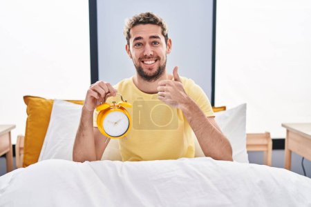 Téléchargez les photos : Homme hispanique avec barbe tenant réveil dans le lit souriant heureux et positif, pouce levé faire excellent et signe d'approbation - en image libre de droit