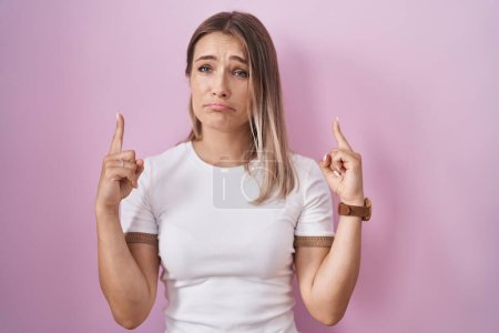 Foto de Blonde caucasian woman standing over pink background pointing up looking sad and upset, indicating direction with fingers, unhappy and depressed. - Imagen libre de derechos