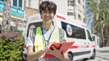 Téléchargez les photos : Jeune homme hispanique infirmière écriture rapport médical debout par ambulance à la rue - en image libre de droit