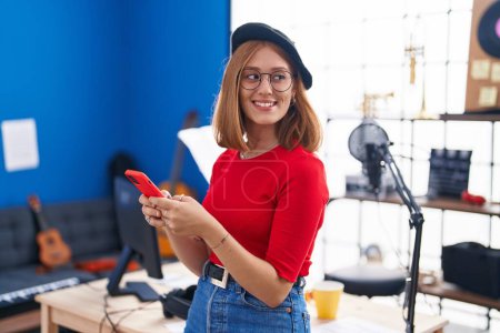 Téléchargez les photos : Jeune rousse femme artiste souriant confiant en utilisant smartphone au studio de musique - en image libre de droit