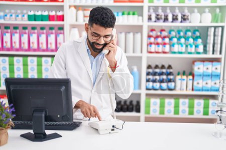Téléchargez les photos : Jeune pharmacien arabe parlant au téléphone à l'aide d'un ordinateur à la pharmacie - en image libre de droit