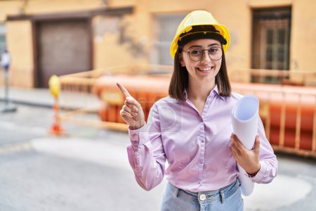 Téléchargez les photos : Fille hispanique portant un casque d'architecte sur le chantier de construction souriant heureux pointant avec la main et le doigt sur le côté - en image libre de droit