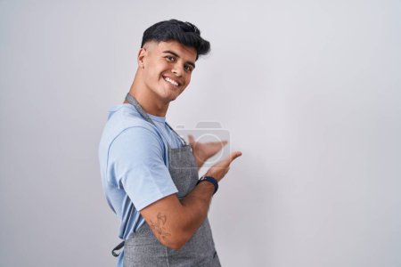Téléchargez les photos : Jeune homme hispanique portant tablier sur fond blanc invitant à entrer souriant naturel avec la main ouverte - en image libre de droit