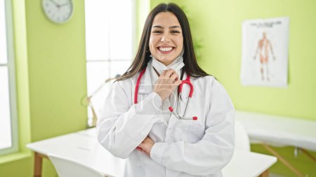 Foto de Joven hermosa mujer hispana médico sacando máscara médica sonriendo en la clínica - Imagen libre de derechos